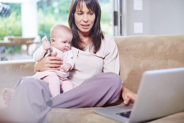 Mother sitting with baby on couch working on laptop - ZEF002282