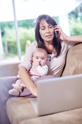 Mutter sitzt mit Baby auf der Couch, arbeitet am Laptop und spricht mit dem Handy - ZEF002283