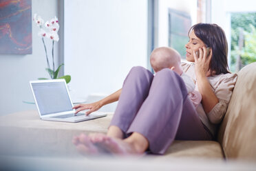 Mother sitting with baby on couch working on laptop and talking on cell phone - ZEF002287