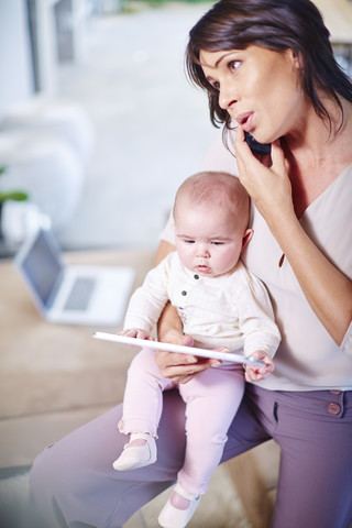 Mutter sitzt mit Baby auf der Couch und spricht mit dem Handy, lizenzfreies Stockfoto