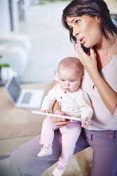 Mother sitting with baby on couch and talking on cell phone - ZEF002289