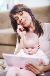 Mother sitting with baby on couch using digital tablet and talking on cell phone - ZEF002291