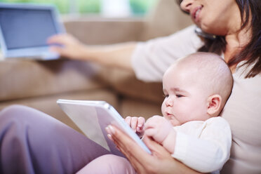 Mutter mit Baby im Wohnzimmer im Gespräch auf dem Handy und Blick auf digitale Tablet - ZEF002292
