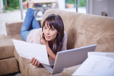 Frau liegt auf der Couch, schaut auf Papier und arbeitet am Laptop - ZEF002270