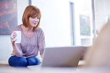 Woman sitting on couch looking at laptop and holding coffee cup - ZEF002280