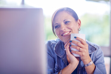 Lächelnde Frau hält eine Kaffeetasse und schaut auf einen Computerbildschirm - ZEF002324