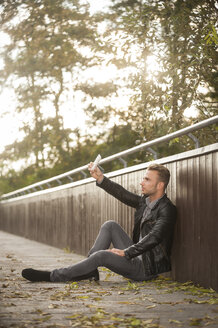 Junger Mann sitzt auf einer Fußgängerbrücke und macht ein Selfie mit seinem Smartphone - PAF001083