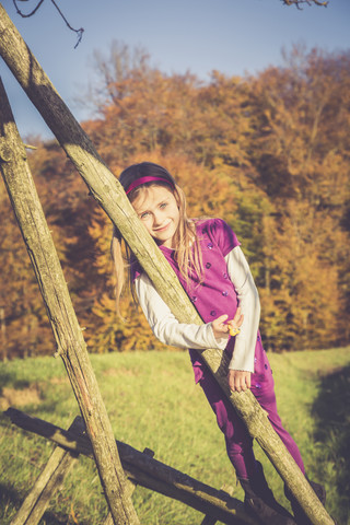 Porträt eines lächelnden kleinen Mädchens, das sich auf eine Holzstange vor einem herbstlichen Wald stützt, lizenzfreies Stockfoto