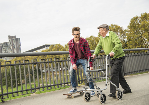 Glücklicher älterer Mann mit Rollator und erwachsenem Enkel mit Skateboard, lizenzfreies Stockfoto