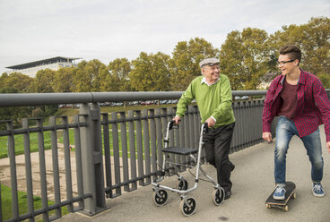 Happy senior man with wheeled walker and adult grandson with skateboard - UUF002651