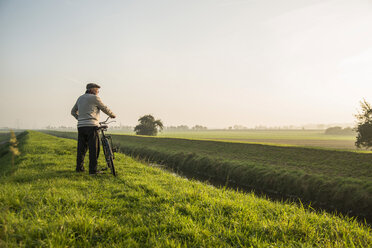 Älterer Mann in ländlicher Landschaft, der ein Fahrrad schiebt - UUF002693