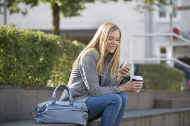 Lächelnde junge Frauen, die auf einer Treppe sitzen und einen Kaffee trinken und dabei ihr Smartphone benutzen - SHKF000031