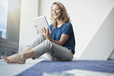 Porträt einer lächelnden reifen Frau, die mit einem digitalen Tablet auf dem Boden in ihrer Wohnung sitzt, lizenzfreies Stockfoto