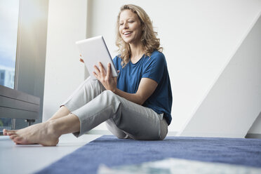 Porträt einer lächelnden reifen Frau, die mit einem digitalen Tablet auf dem Boden in ihrer Wohnung sitzt - RBF002033