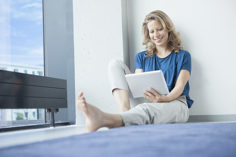 Porträt einer lächelnden reifen Frau, die mit einem digitalen Tablet auf dem Boden in ihrer Wohnung sitzt, lizenzfreies Stockfoto