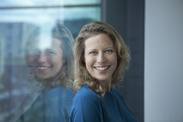Porträt einer lächelnden reifen Frau, die sich am Fenster ihrer Wohnung anlehnt - RBF002040