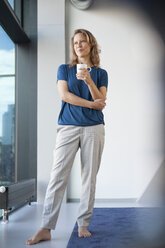 Smiling mature woman with mug looking through window at her apartment - RBF002042