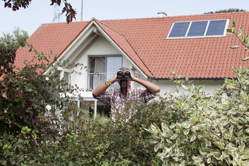 Mann schaut durch ein Fernglas über eine Hecke - RBF001904