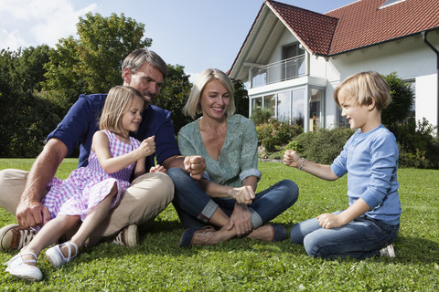 Glückliche Familie sitzt auf dem Rasen im Garten und spielt Stein-Papier-Schere, lizenzfreies Stockfoto