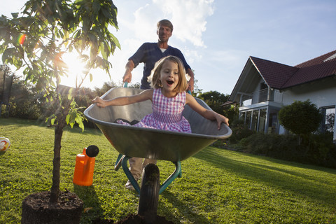 Vater mit Tochter in Schubkarre pflanzt Baum im Garten, lizenzfreies Stockfoto