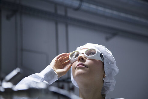 Junge Technikerin mit Schutzkappe und Schutzbrille schaut in einem Laboratorium nach oben - SGF001167