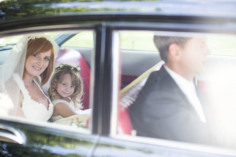 Braut sitzt im Auto mit kleiner Brautjungfer und Fahrer vor der Hochzeit, lizenzfreies Stockfoto