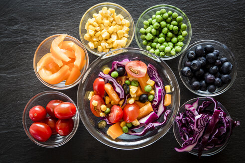 Gemischter Salat und Glasschalen mit verschiedenen Rohkostsorten in Regenbogenfarben auf Schiefer angeordnet - SARF001060