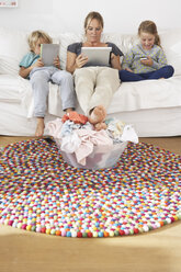 Mother and two children sitting on couch using smartphone and digital tablets ignoring pile of laundry - FSF000289