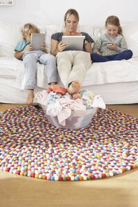 Mutter und zwei Kinder sitzen auf der Couch und nutzen Smartphone und digitale Tablets, während sie einen Haufen Wäsche ignorieren - FSF000289