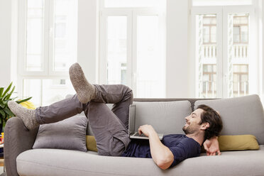 Relaxed man lying on couch using laptop - FMKF001387