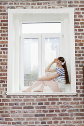 Glückliche Frau sitzt auf der Fensterbank und hört Musik - FMKF001399