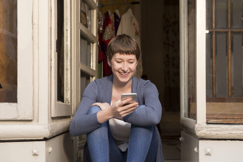 Portrait of female fashion designer with smartphone sitting at the entrance of her studio - DISF001114