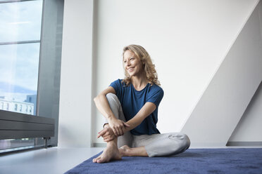 Portrait of happy mature woman sitting on carpet at home looking through window - RBF002056