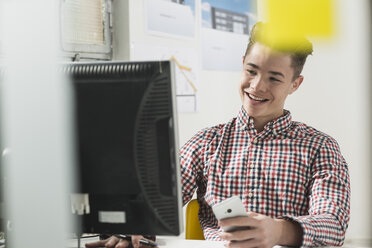 Smiling young man at desk with computer and cell phone - UUF002855