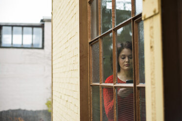 Junge Frau mit Mobiltelefon hinter dem Fenster - UUF002874