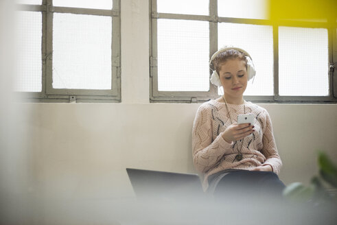 Junge Frau sitzt mit Kopfhörern und Handy auf dem Schreibtisch - UUF002935
