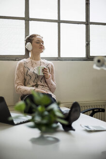 Junge Frau sitzt mit Kopfhörern und einer Tasse Kaffee am Schreibtisch - UUF002936