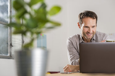 Lächelnder Geschäftsmann mit Blick auf den Laptop im Büro - UUF002942