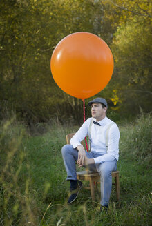 Man sitting on chair with balloon on meadow - JTLF000018