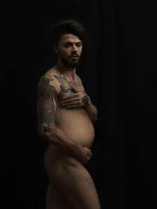 Nackter schwangerer Mann mit Tatoos vor schwarzem Hintergrund - STK001130