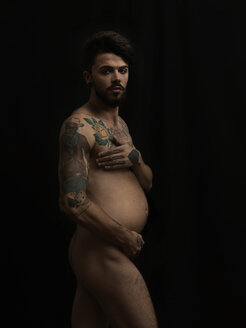 Naked pregnant man with tatoos in front of black background - STK001130