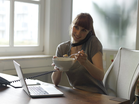 Lächelnde Frau mit Laptop am Schreibtisch, die telefoniert und aus einer Schüssel isst, lizenzfreies Stockfoto