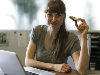 Portrait of woman at desk with laptop eating bread - STKF001191