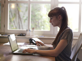 Woman at desk using laptop and eating muesli - STKF001157