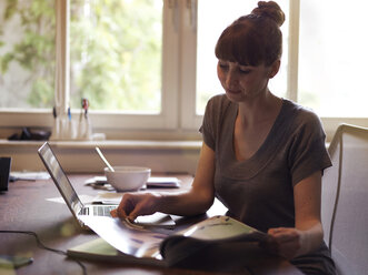 Frau am Schreibtisch mit Laptop, die eine Zeitschrift liest - STKF001193