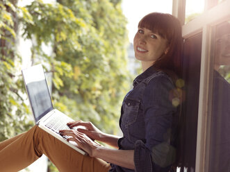 Lächelnde Frau am offenen Fenster mit Laptop - STKF001163