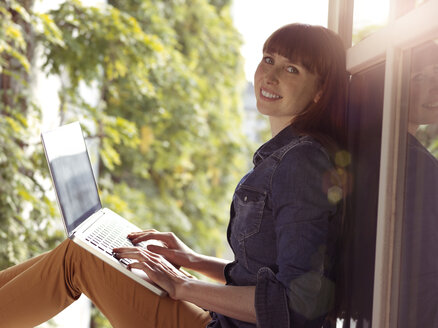 Smiling woman at open window using laptop - STKF001163