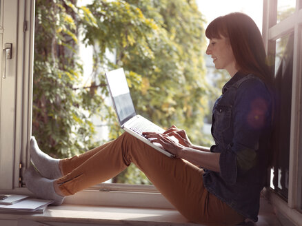 Smiling woman at open window using laptop - STKF001182