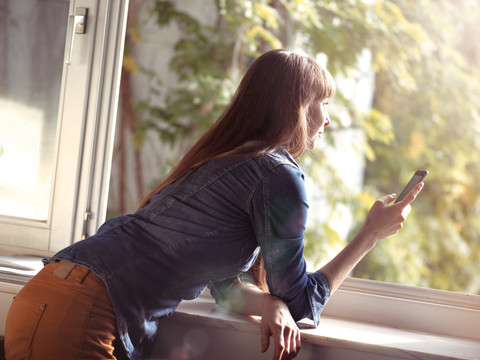 Frau am offenen Fenster mit Handy in der Hand, lizenzfreies Stockfoto