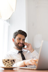 Businessman with laptop and cup of coffee on cell phone - WESTF020479